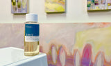 A bottle of Gamblin 3-in-1 oil painting medium on display in front of pastel and yellow abstract surfboard paintings