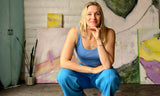 San Diego artist Skye Silver squatting down and posing in front of her colorful abstract surf series paintings, a snake plant, and Chris Rudy surfboard