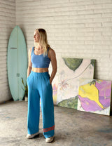 artist Skye Silver standing in her art studio wearing an athletic blue track suit with her abstract beach culture paintings and a blue surfboard in the background